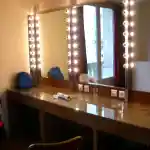 Platée - Opéra-Comique de Paris | La photographie que j’ai prise capture l’intérieur du côté droit de la loge de l’Opéra Comique à Paris. La pièce est éclairée par de grosses ampoules rondes aux tons jaunes, encadrant trois grands miroirs muraux, créant une ambiance chaleureuse. Le meuble en bois, sur lequel repose une épaisse plaque de verre transparente servant de coiffeuse, est agencé de manière à offrir un espace de préparation optimal pour les artistes. Sur ce dernier, se trouvent trois verres en plastique remplis d’eau ainsi qu'un téléphone portable et contre le mur du fond, un petit sac à dos bleu.