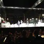 Platée - Opéra-Comique de Paris | Cette image capture un moment magique au sein d’un opéra comique, où les artistes sur scène sont éclairés, contrastant avec l’obscurité de l’orchestre et du public en premier plan. Le décor élégant et les costumes des artistes évoquent une performance sophistiquée et captivante.